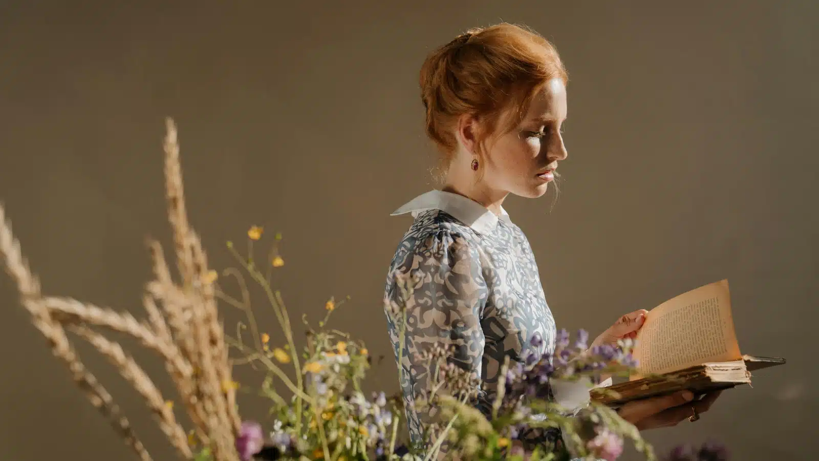Image for the "women's impact on film fashion and fiction" article, a side view of a red headed woman in a floral dress reading a book, with a dried flower arrangement in the foreground